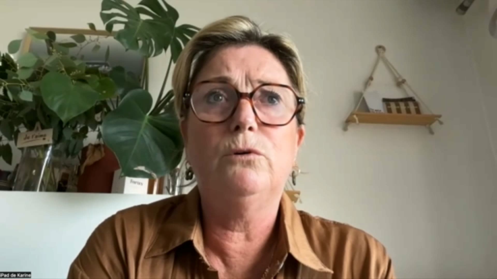 Portrait d’une femme assise en intérieur, portant des lunettes, avec des plantes en arrière-plan et un mur clair décoré d’une petite étagère.