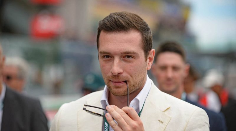 Homme jeune portant une veste claire tenant une paire de lunettes devant lui dans un environnement extérieur animé.