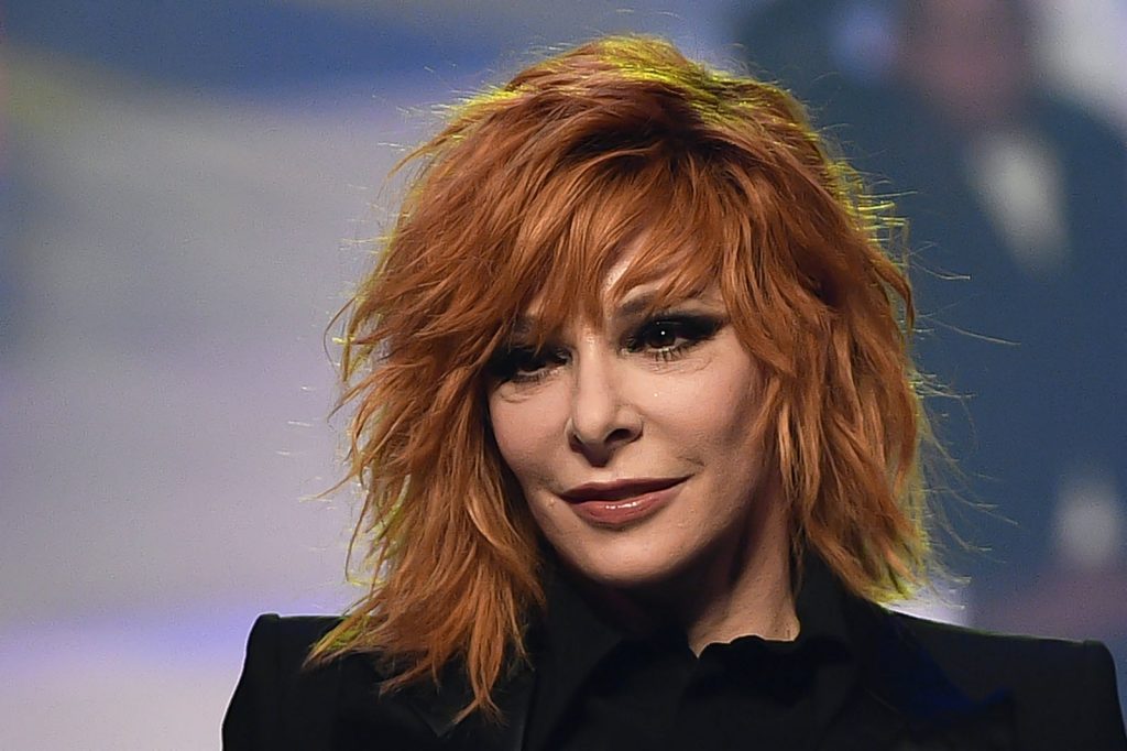 Portrait d’une femme rousse sous un éclairage de scène, avec un maquillage prononcé et un fond flou.