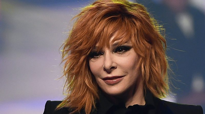 Portrait d’une femme rousse sous un éclairage de scène, avec un maquillage prononcé et un fond flou.