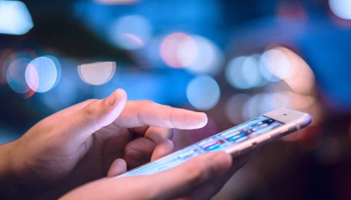Main tenant un smartphone allumé, doigt en train de toucher l’écran, utilisation d’un téléphone mobile dans un environnement urbain flou et lumineux