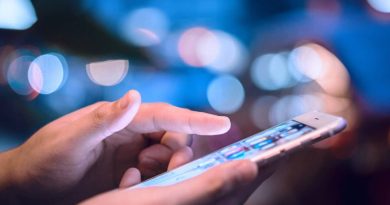 Main tenant un smartphone allumé, doigt en train de toucher l’écran, utilisation d’un téléphone mobile dans un environnement urbain flou et lumineux