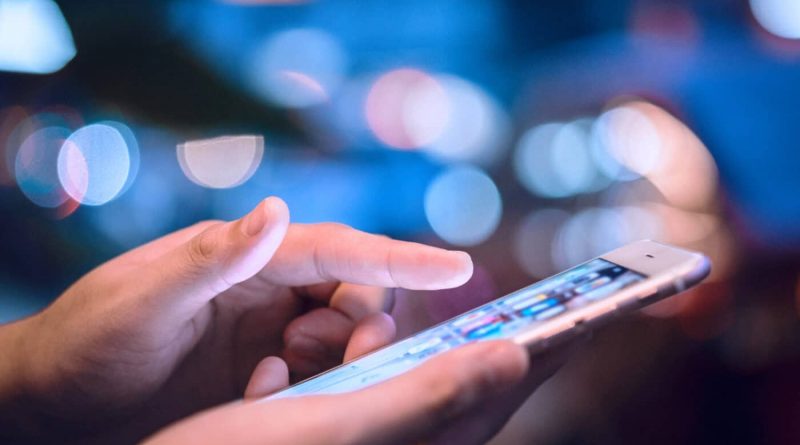 Main tenant un smartphone allumé, doigt en train de toucher l’écran, utilisation d’un téléphone mobile dans un environnement urbain flou et lumineux