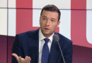 Homme politique en costume sombre s’exprimant sur un plateau de télévision lors d’un débat en direct, avec un geste de la main et une expression surprise