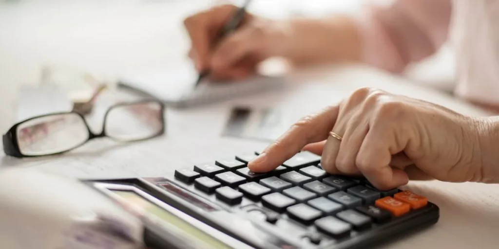 Main d’une personne âgée utilisant une calculatrice sur une table avec des documents et des lunettes posées à côté.