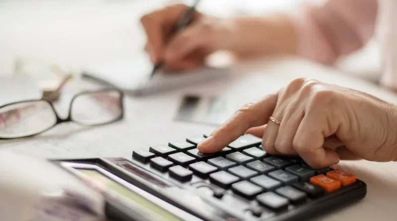 Main d’une personne âgée utilisant une calculatrice sur une table avec des documents et des lunettes posées à côté.