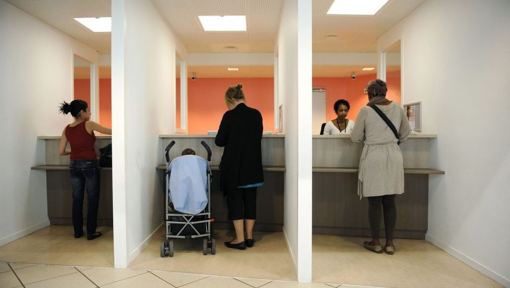Personnes debout devant des guichets dans un service administratif, avec une poussette au centre.