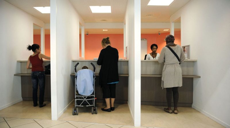 Personnes debout devant des guichets dans un service administratif, avec une poussette au centre.