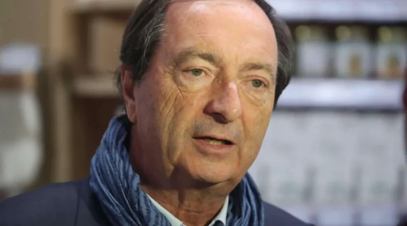 Portrait en gros plan d’un homme d’âge mûr portant une veste sombre et une écharpe bleue, filmé lors d’une interview, avec un arrière-plan flou évoquant un intérieur avec des étagères et des objets.