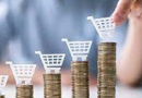 Main tenant un chariot de courses miniature posé sur des piles de pièces de monnaie de hauteurs croissantes, illustrant l’écart entre le travail fourni et le pouvoir d’achat réel