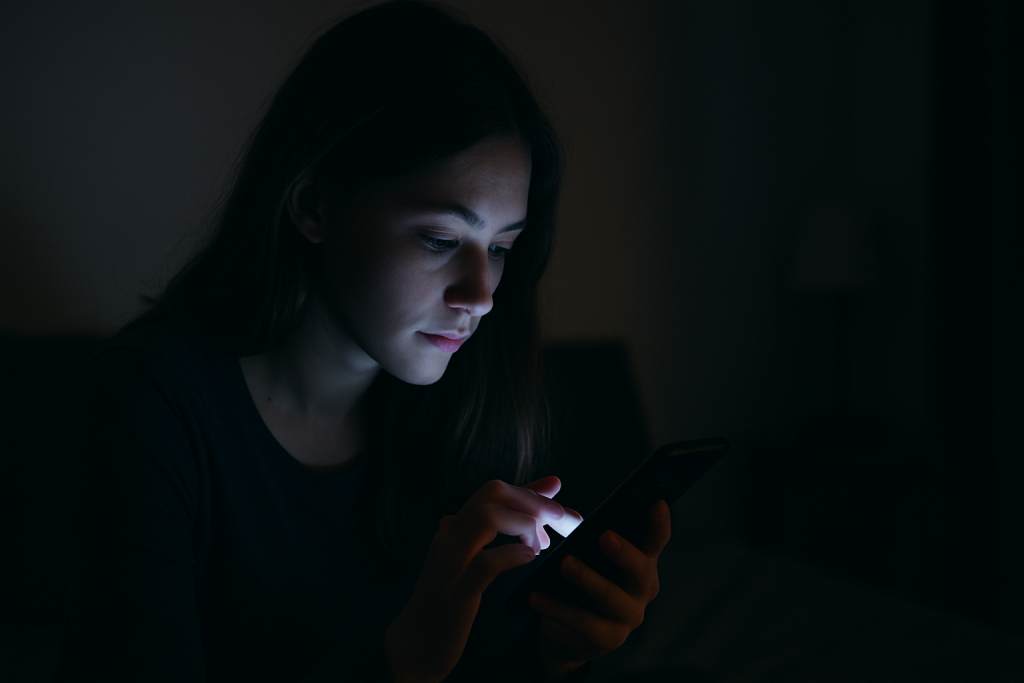 Personne seule utilisant un smartphone dans l’obscurité, visage éclairé par la lumière de l’écran dans une ambiance nocturne intime