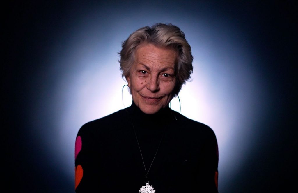Portrait d’une femme mature éclairée par un halo lumineux, vêtue d’un haut sombre, regard direct et expression douce.