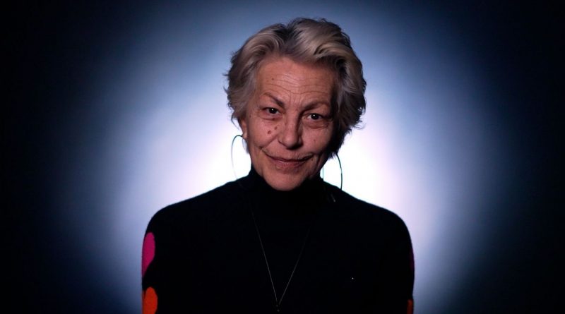 Portrait d’une femme mature éclairée par un halo lumineux, vêtue d’un haut sombre, regard direct et expression douce.