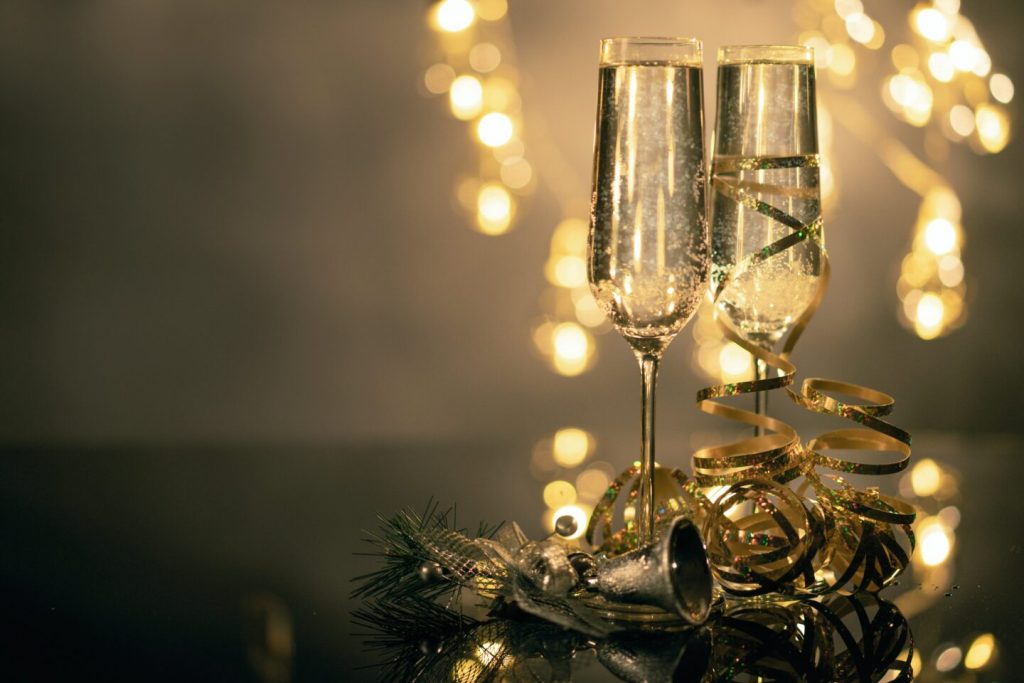 Deux flûtes de fête remplies d’une boisson pétillante, sur une table sombre, entourées de décorations dorées et de lumières floues en arrière-plan.