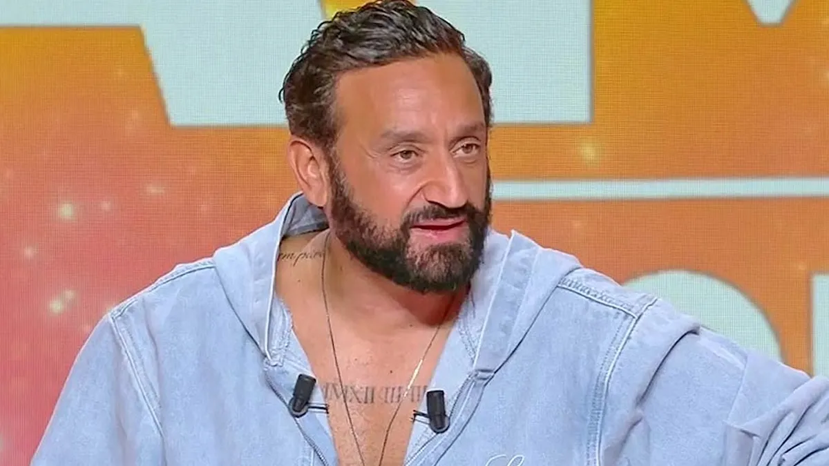 Animateur barbu en gros plan sur un plateau TV, parlant face caméra, avec un fond orange lumineux derrière lui.