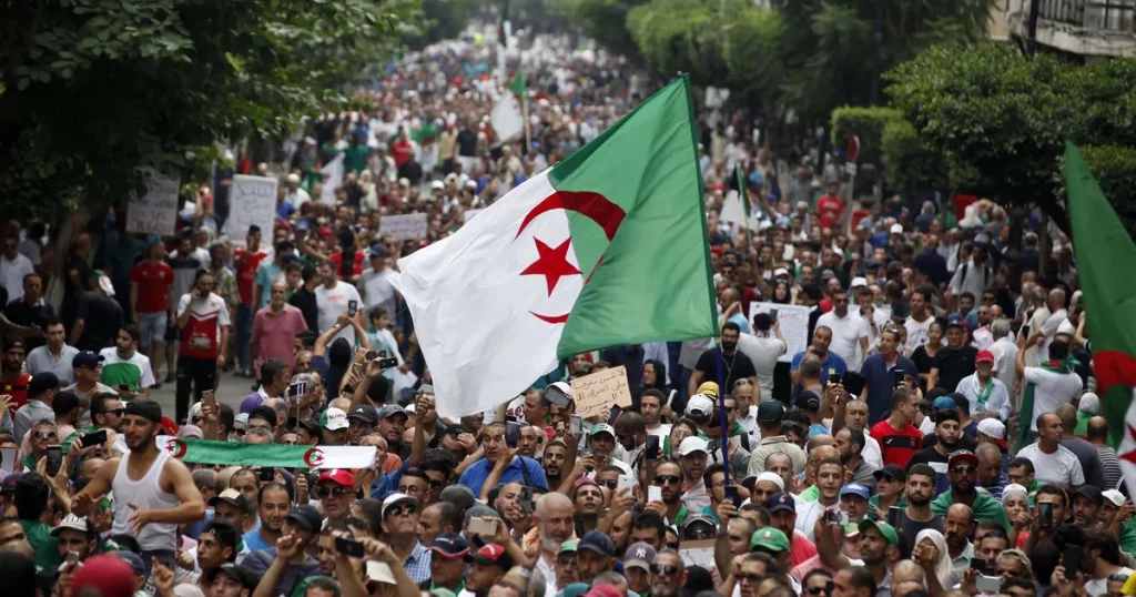 Grande foule rassemblée lors d’une manifestation, avec des drapeaux vert, blanc et rouge brandis au-dessus des participants dans une avenue bordée d’arbres.