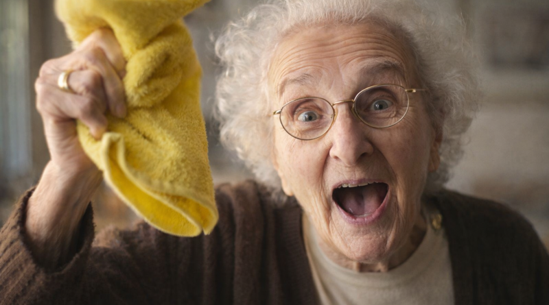 Femme âgée souriante brandissant un chiffon jaune, en train de faire le ménage à l’intérieur d’une maison.