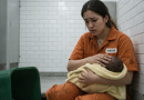 Jeune femme assise au sol dans une pièce carrelée, tenant un nouveau-né emmailloté contre elle, avec une poubelle à proximité, dans une atmosphère tendue et dramatique.