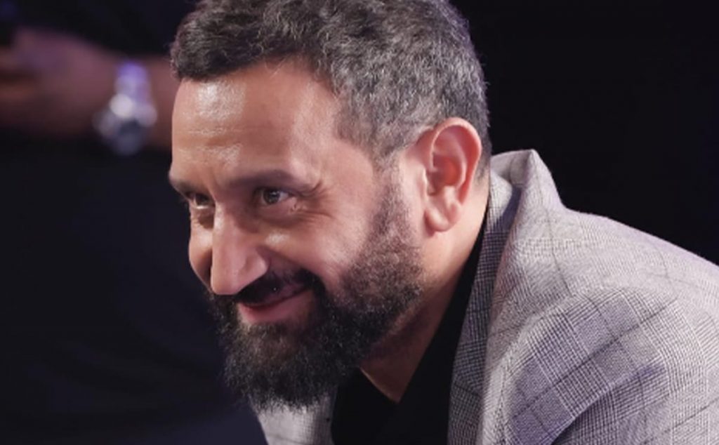 Portrait rapproché d’un animateur de télévision barbu, souriant légèrement, vêtu d’une veste claire, dans une ambiance de studio sombre et feutrée.