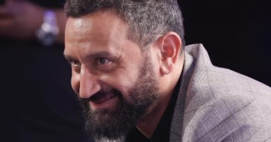 Portrait rapproché d’un animateur de télévision barbu, souriant légèrement, vêtu d’une veste claire, dans une ambiance de studio sombre et feutrée.