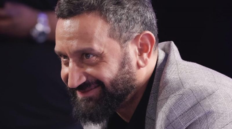 Portrait rapproché d’un animateur de télévision barbu, souriant légèrement, vêtu d’une veste claire, dans une ambiance de studio sombre et feutrée.