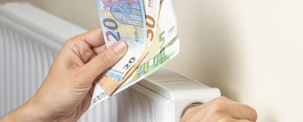 Main tenant des billets en euros devant un radiateur domestique, symbolisant le lien entre chauffage, consommation d’énergie et budget du foyer.