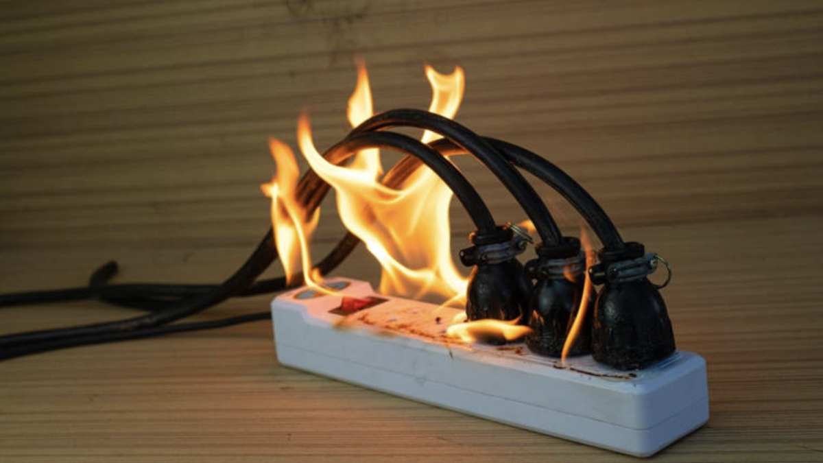 Multiprise électrique posée sur une surface en bois, avec plusieurs câbles branchés en surchauffe et des flammes visibles autour des prises.
