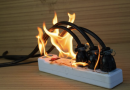 Multiprise électrique posée sur une surface en bois, avec plusieurs câbles branchés en surchauffe et des flammes visibles autour des prises.