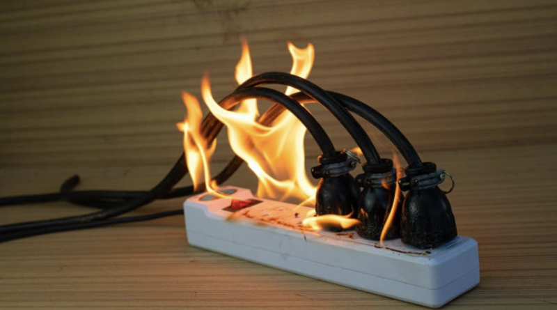 Multiprise électrique posée sur une surface en bois, avec plusieurs câbles branchés en surchauffe et des flammes visibles autour des prises.