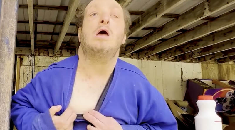 Personne adulte assise dans un abri en bois, portant un gilet bleu entrouvert, regard levé, environnement rustique et désordonné en arrière-plan.