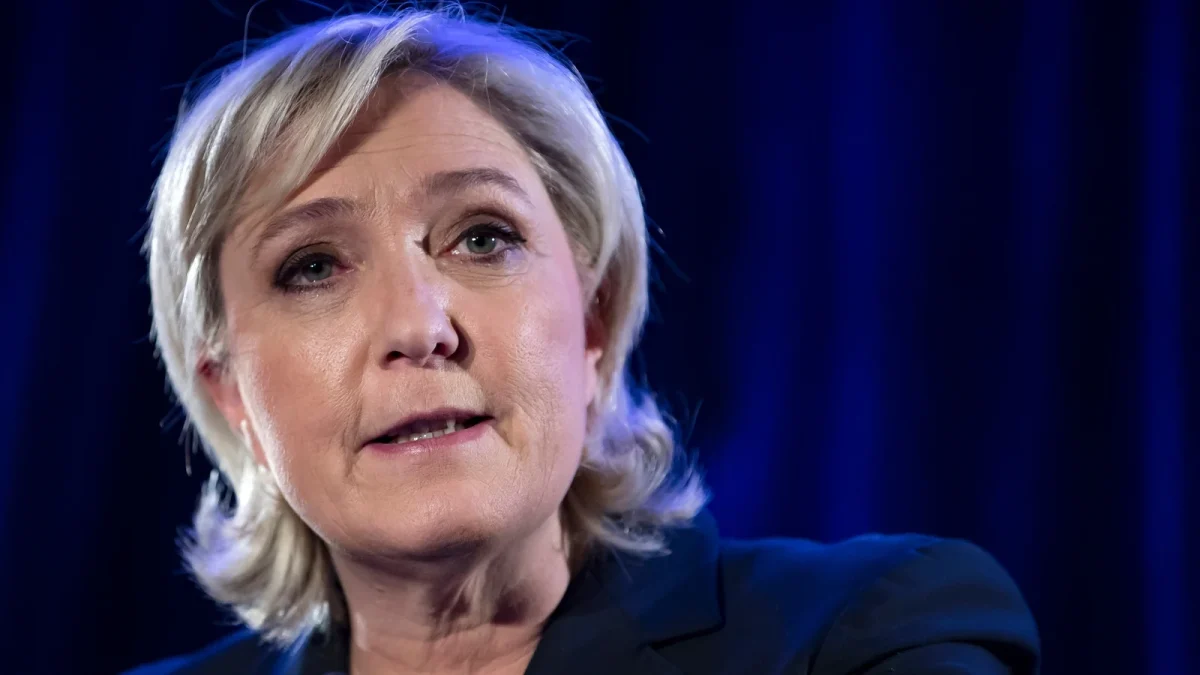 Portrait serré d’une femme politique lors d’une prise de parole publique, éclairage bleu en arrière-plan, expression sérieuse et concentrée, ambiance institutionnelle.