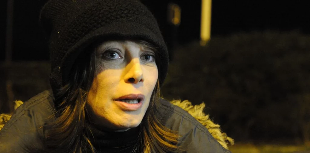 gros plan nocturne d’une femme emmitouflée dans un manteau et un bonnet, le regard inquiet éclairé par un lampadaire