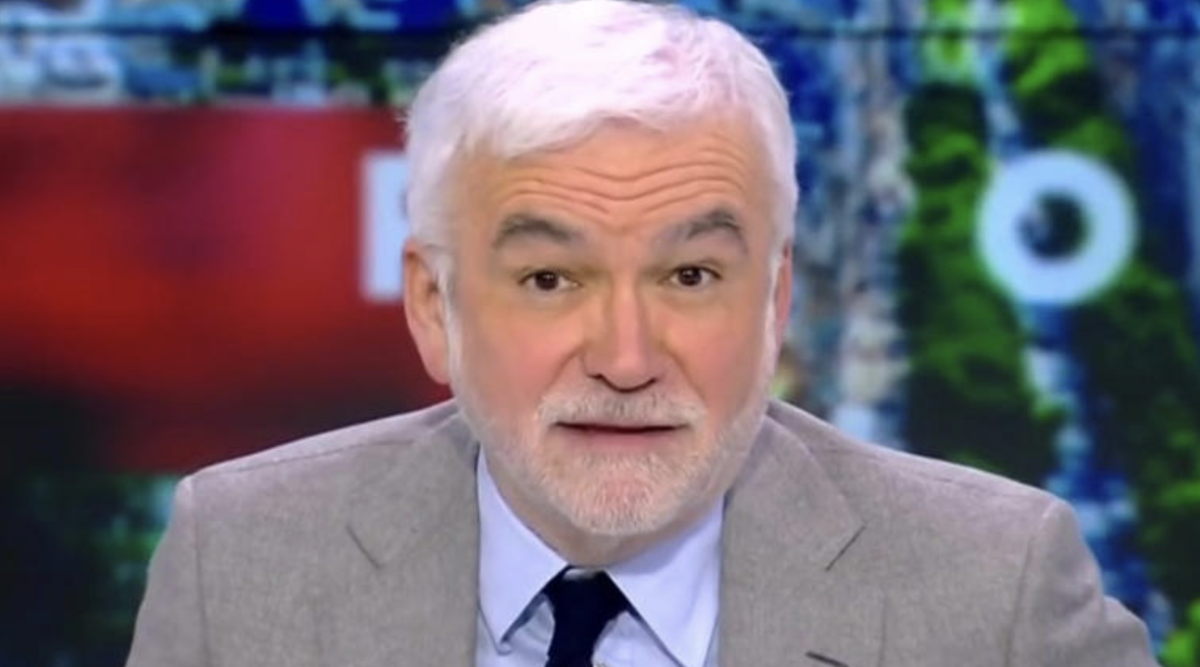 Présentateur de télévision aux cheveux blancs et à la barbe claire, regard face caméra, assis en plateau dans un studio d’actualité aux couleurs contrastées.