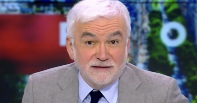 Présentateur de télévision aux cheveux blancs et à la barbe claire, regard face caméra, assis en plateau dans un studio d’actualité aux couleurs contrastées.