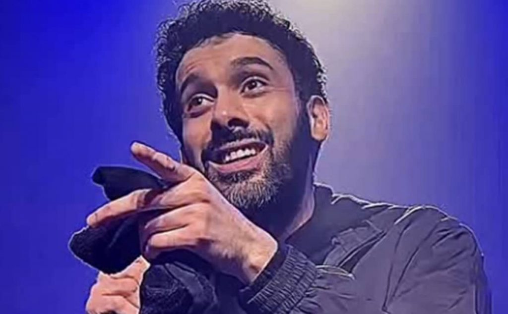 Humoriste sur scène tenant un micro, sourire ironique, geste de la main vers le public, éclairage bleu de spectacle, ambiance de stand-up.