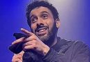 Humoriste sur scène tenant un micro, sourire ironique, geste de la main vers le public, éclairage bleu de spectacle, ambiance de stand-up.