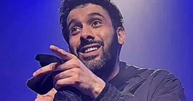 Humoriste sur scène tenant un micro, sourire ironique, geste de la main vers le public, éclairage bleu de spectacle, ambiance de stand-up.