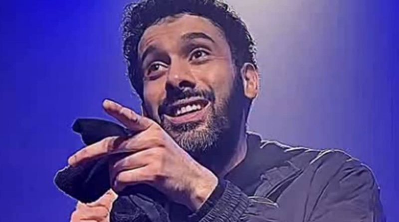 Humoriste sur scène tenant un micro, sourire ironique, geste de la main vers le public, éclairage bleu de spectacle, ambiance de stand-up.