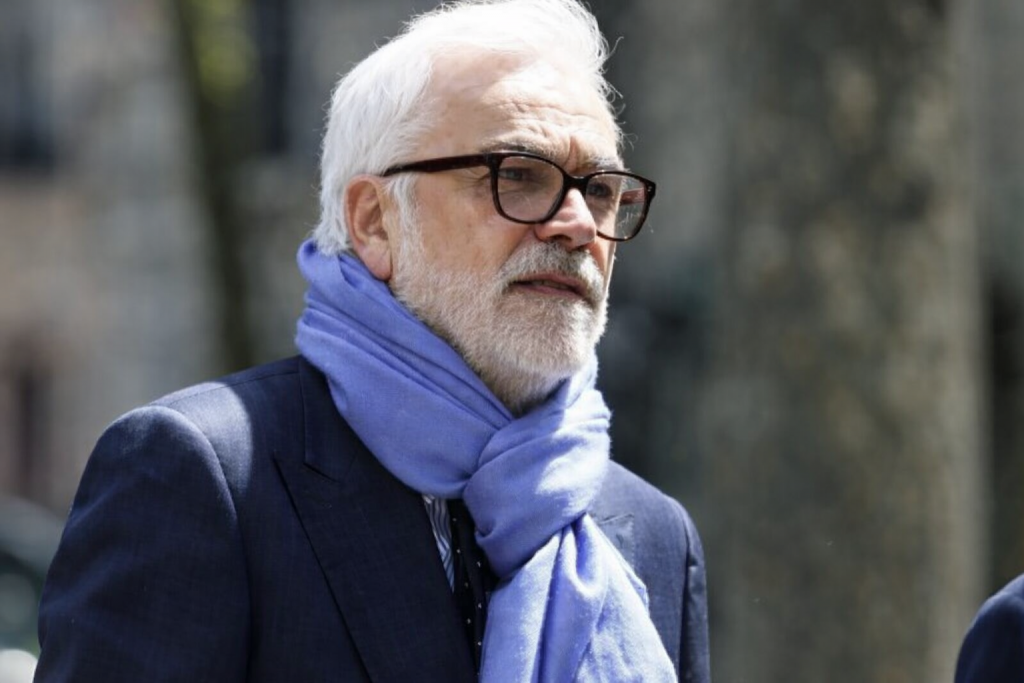 Homme mûr aux cheveux blancs et barbe soignée, portant des lunettes et une écharpe claire, photographié en extérieur dans un environnement urbain flouté
