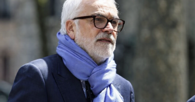 Homme mûr aux cheveux blancs et barbe soignée, portant des lunettes et une écharpe claire, photographié en extérieur dans un environnement urbain flouté