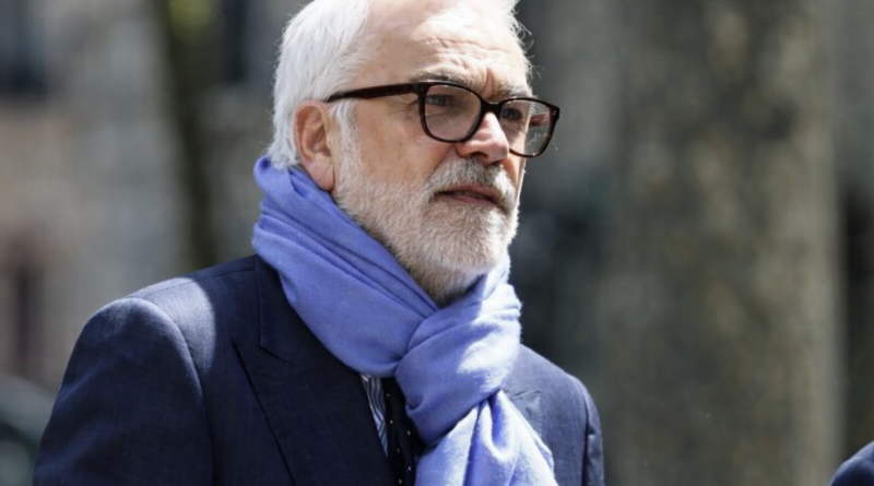 Homme mûr aux cheveux blancs et barbe soignée, portant des lunettes et une écharpe claire, photographié en extérieur dans un environnement urbain flouté