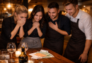 Groupe de serveurs réunis autour d’une table en bois, visiblement émus et souriants, observant une importante somme d’argent déposée devant eux dans un restaurant à l’ambiance chaleureuse.