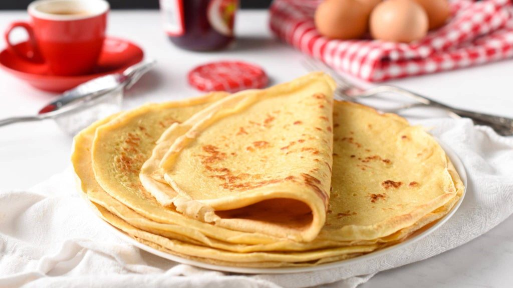 Crêpes fines et dorées empilées dans une assiette blanche, posées sur une table lumineuse avec des œufs et une tasse en arrière-plan, évoquant une tradition gourmande et conviviale de l’hiver.