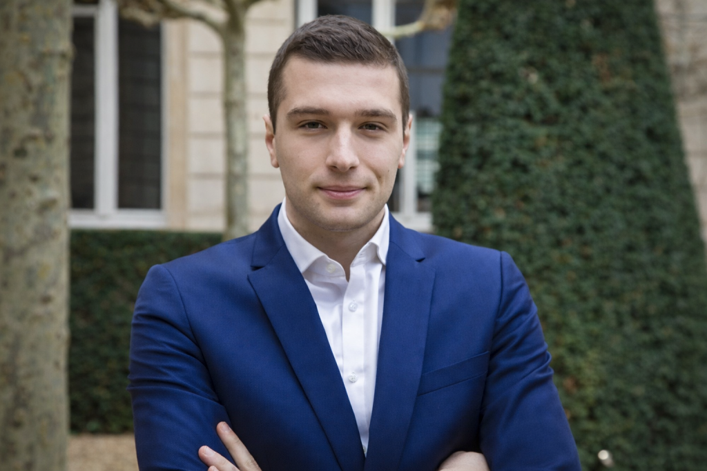 Homme politique en costume bleu foncé, bras croisés, photographié en extérieur devant un bâtiment institutionnel et de la végétation