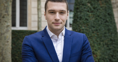 Homme politique en costume bleu foncé, bras croisés, photographié en extérieur devant un bâtiment institutionnel et de la végétation