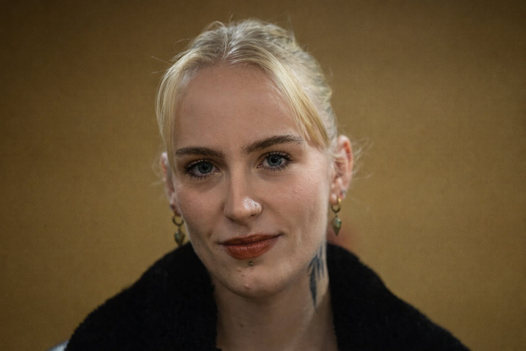 Portrait serré d’une jeune femme blonde regardant l’objectif, expression calme et déterminée, fond flou aux tons chauds, ambiance sobre et journalistique.