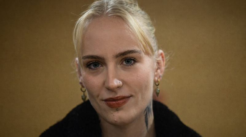 Portrait serré d’une jeune femme blonde regardant l’objectif, expression calme et déterminée, fond flou aux tons chauds, ambiance sobre et journalistique.