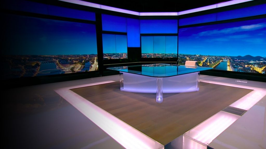 Plateau de télévision vide, éclairé par des lumières bleutées et violacées, avec un bureau central moderne et des écrans panoramiques diffusant une vue urbaine nocturne en arrière-plan, créant une atmosphère froide et institutionnelle.