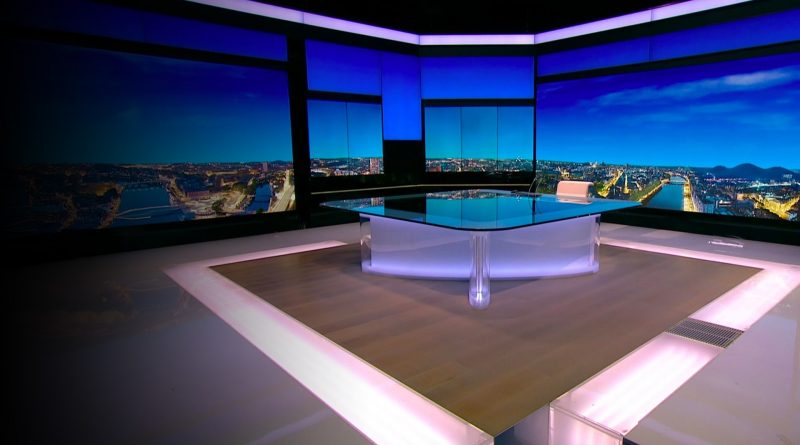 Plateau de télévision vide, éclairé par des lumières bleutées et violacées, avec un bureau central moderne et des écrans panoramiques diffusant une vue urbaine nocturne en arrière-plan, créant une atmosphère froide et institutionnelle.