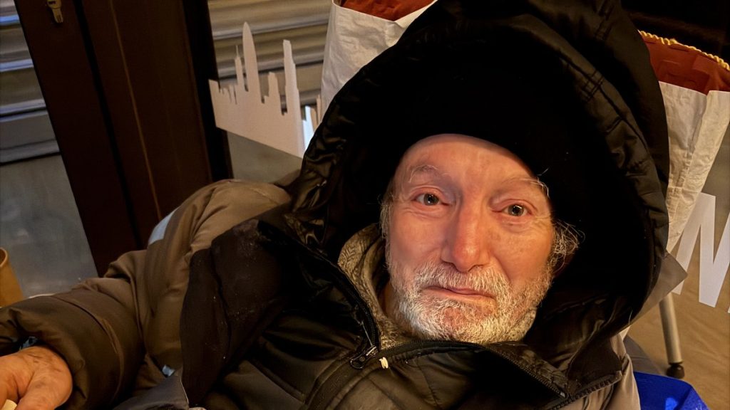 Homme âgé emmitouflé dans un manteau épais, assis à l’intérieur d’un abri urbain, le visage marqué par la fatigue et le froid, regard humide et épuisé.